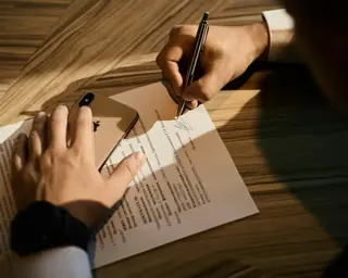 Hands completing a printed form on a wooden desk in raking light