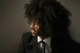 A person in a dark suit and tie photographed against a dark ground