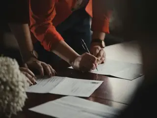 Two people signing a document across a table under a single warm light
